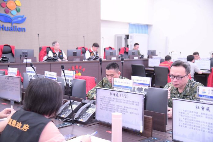 Military briefing room with soldiers at desks, monitors, and microphones; officials sit at a long table in the background.